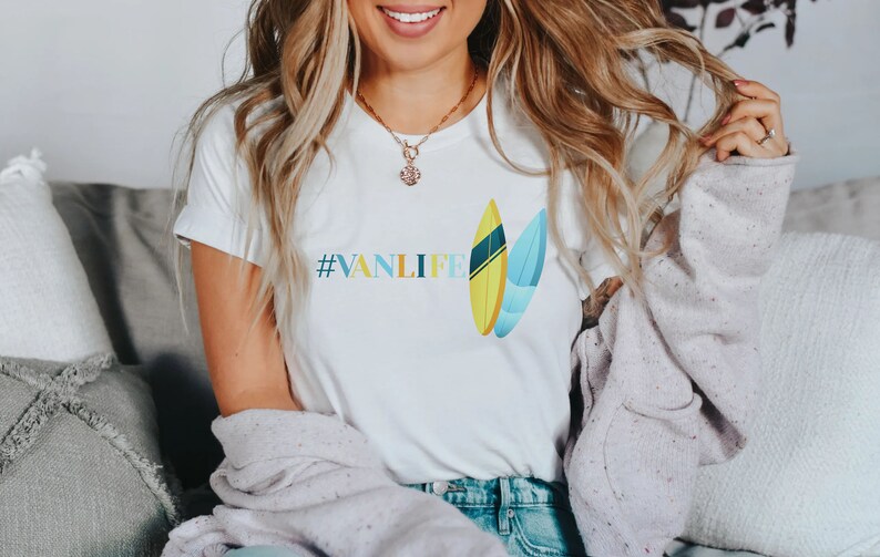 Peut inclure: T-shirt blanc avec le texte "#VANLIFE" et un graphique de deux planches de surf. Les planches de surf sont jaunes, bleues et vertes.