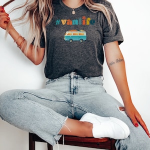 Puede incluir: Una mujer lleva una camiseta gris oscuro con el texto "#vanlife" y una ilustraci&oacute;n retro de una furgoneta azul. La camiseta se combina con unos vaqueros azules claros y zapatillas blancas.