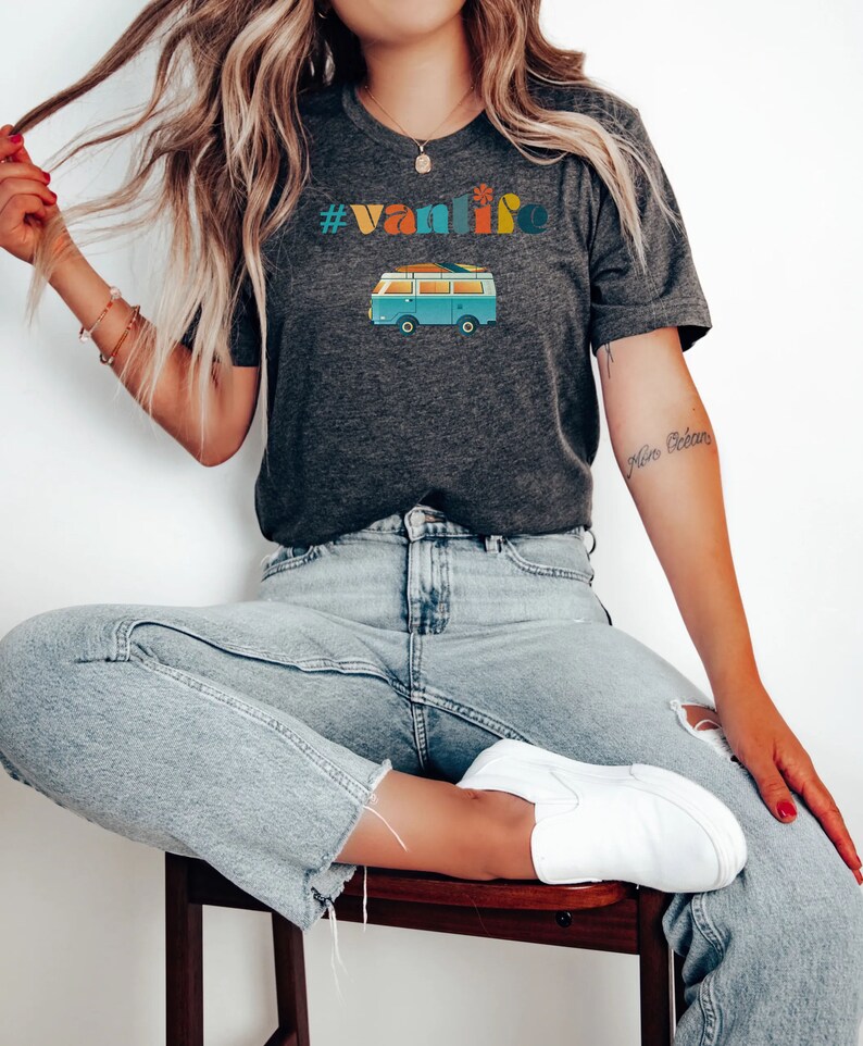 Puede incluir: Una mujer lleva una camiseta gris oscuro con el texto "#vanlife" y una ilustraci&oacute;n retro de una furgoneta azul. La camiseta se combina con unos vaqueros azules claros y zapatillas blancas.