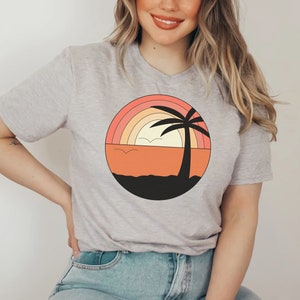 Puede incluir: Una camiseta gris con un gráfico de una palmera y una puesta de sol con un arcoíris. El gráfico está en tonos de naranja, rosa, amarillo y negro.