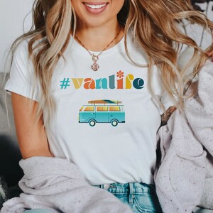 Puede incluir: Camiseta blanca con el texto "#vanlife" en letras de colores y una furgoneta vintage azul con una tabla de surf en la parte superior.