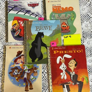 May include: A collection of Disney Pixar Little Golden Books. Titles include "Cars", "Finding Nemo", "Brave", and "Presto!". The books feature colorful illustrations and gold-colored spines. The books are arranged on a white lace surface.