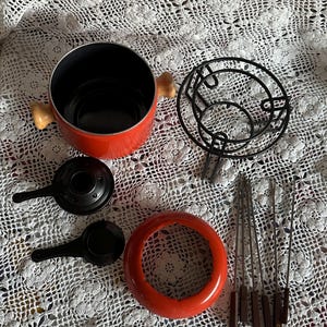 May include: A fondue set featuring a bright orange pot with light wood handles, a black metal stand, a black burner with a lid, a matching orange base ring, and six metal fondue forks with brown square handles arranged on a white lace cloth.