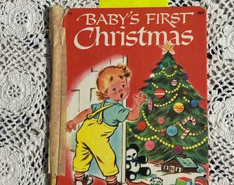 May include: Vintage children's book titled "Baby's First Christmas" with a red cover. The cover illustration features a young child in yellow overalls and a blue shirt, standing near a decorated Christmas tree. The book has a worn appearance.