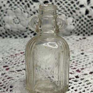 May include: A small, clear glass bottle featuring a narrow neck with two integrated loop handles on the sides. The body of the bottle has a vertical ribbed texture and a flat base, creating a distinct silhouette with a transparent finish.