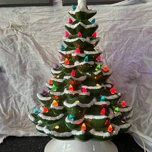Op de afbeelding: Een groene keramische kerstboom met een witte, sneeuwachtige textuur op de takken, verlicht door kleine, veelkleurige plastic lampjes in rood, blauw, oranje en roze, geplaatst op een ronde witte voet met een bijbehorend elektrisch snoer.
