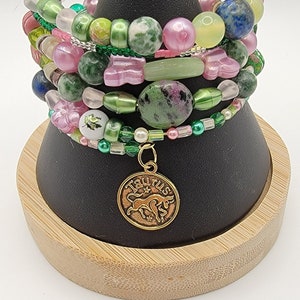 May include: A set of six green, pink, and white beaded bracelets with a gold Taurus zodiac charm. The bracelets are stacked on a black display stand.