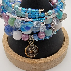 May include: A set of six colorful beaded bracelets with a gold charm featuring a crab. The bracelets are stacked on a wooden display stand.