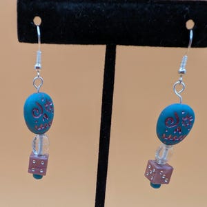 Può includere: Un paio di orecchini pendenti. Ogni orecchino presenta una perla ovale color teal con un disegno floreale rosso, una perla trasparente e una perla a cubo marrone. Gli orecchini pendono da ganci color argento.