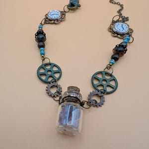 Puede incluir: Un collar de estilo steampunk con un pequeño dije de botella de vidrio, dos caras de reloj antiguas y engranajes de bronce. El collar está hecho con cuentas marrones y turquesas y una cadena de bronce.
