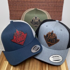 May include: Three trucker hats with a mesh back. The hats are in different colors: navy blue, gray, and green. Each hat has a patch on the front that says "Top Dad" with a star and wings.
