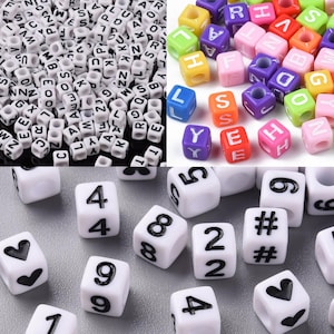 Könnte beinhalten: Eine Sammlung weißer quadratischer Perlen mit schwarzen Buchstaben, Zahlen und Symbolen. Einige Perlen sind farbig mit weißen Buchstaben. Geeignet für Schmuckherstellung und Bastelarbeiten.