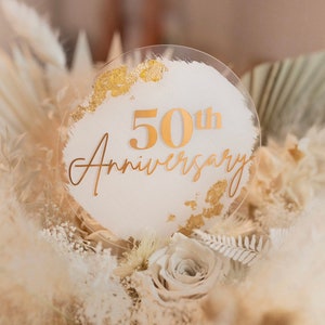 以下が含まれることがあります： 「50th Anniversary」と金色の文字が書かれた白いアクリル製の丸い看板。看板は金色のグリッターで飾られており、白いパンパスグラスのベッドの上に置かれています。