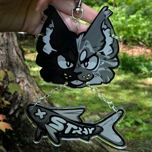 May include: A clear acrylic keychain featuring a split-faced cat design in black and gray, connected by a chain to a gray fish with the word "STRAY" in white. The keychain has a silver clasp.