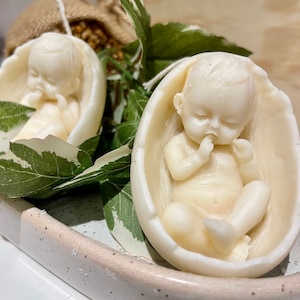 May include: Two white baby candles in egg-shaped holders. The candles are sculpted to look like sleeping babies. The candles are on a white tray with green leaves.