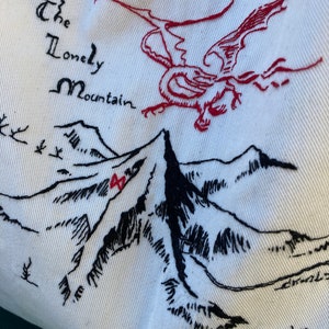 以下が含まれることがあります： 「The Lonely Mountain」という文字と、その上を飛ぶ赤いドラゴンが刺繍された山脈のデザイン。白い布地に刺繍されています。