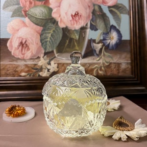 May include: A clear glass jar with a lid, featuring a cut-glass design. The jar is filled with a white substance and has a light yellow hue. The jar is sitting on a light brown surface.