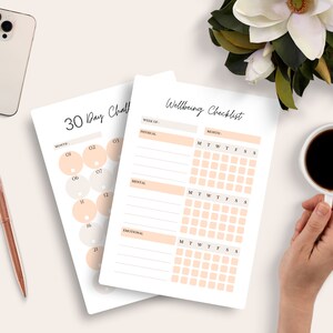 May include: Two white paper checklists, one labeled "30 Day Challenge" and the other "Wellbeing Checklist", are displayed on a light surface. The "30 Day Challenge" checklist has numbered circles. The "Wellbeing Checklist" has sections for physical, mental, and emotional well-being.