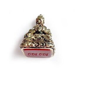 May include: An antique gold-coloured seal with intricate detailing. The seal has a rectangular, red stone base with engraved text. The top of the seal features a decorative floral and scroll design. A unique vintage item.