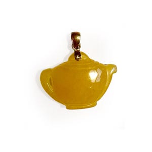 May include: A yellow, translucent, teapot-shaped pendant carved from stone. The pendant features a smooth, polished surface and is attached to a simple, metallic gold-toned bail at the top, designed for suspension on a necklace chain or cord.
