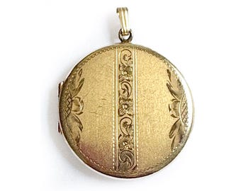 May include: A round, gold-colored locket with a decorative floral and striped design. The locket has a hinged opening and a small loop at the top for a chain. The surface has a slightly aged appearance.
