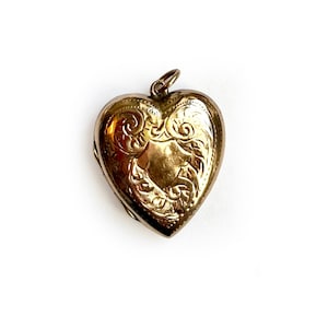 Puede incluir: Un relicario dorado en forma de corazón con intrincados grabados florales y de volutas. El relicario tiene un pequeño lazo en la parte superior para una cadena. La joya vintage está sobre un fondo blanco. Un recuerdo romántico.