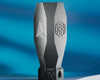 Può includere: Un oggetto geometrico grigio scuro sfaccettato con una superficie testurizzata. Presenta un emblema triangolare su un lato e un motivo geometrico circolare sull'altro. La base è nera, contrassegnata con il testo GAUSS 1100D e un pulsante.