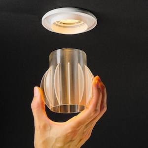 Könnte beinhalten: Ein transparenter, zylindrischer Lichtdiffusor mit vertikalen Lamellen, der für die Befestigung an einer Einbaudeckenleuchte vorgesehen ist. Das durchscheinende, gerippte Kunststoffteil hat eine hohle Mitte und eine breite, runde Öffnung.