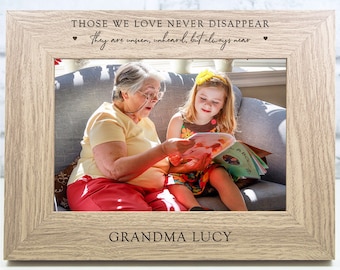 May include: A wooden photo frame with a light brown finish. The frame has a white mat and a photo of an older woman and a young girl reading a book. The frame has the text "THOSE WE LOVE NEVER DISAPPEAR they are unseen, unheard, but always near" and "GRANDMA LUCY" engraved on it.