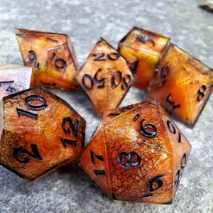 Peut inclure: Un ensemble de dés polyédriques avec un effet marbré ambre et brun. Les dés ont des chiffres noirs et sont conçus pour les jeux de rôle sur table. Les dés sont présentés sur une surface grise texturée.