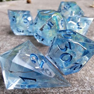 Peut inclure: Un ensemble de sept dés polyédriques bleus, avec des paillettes incrustées et des chiffres bleu foncé. Les dés transparents révèlent des paillettes et des tourbillons internes. Ces dés sont conçus pour les jeux de rôle sur table.