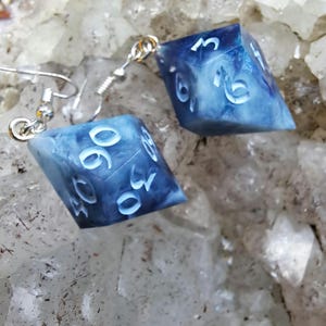 May include: A pair of dice-shaped earrings. Each earring is a blue and white marbled cube with white numbers. The earrings hang from silver-colored ear wires. The earrings are displayed on a white and gray surface.