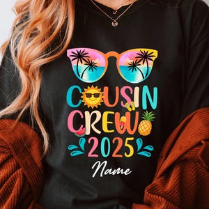 May include: Black t-shirt with a colorful graphic design featuring sunglasses, a sun, watermelon, pineapple, and the text "Cousin Crew 2025" with a space for a name.