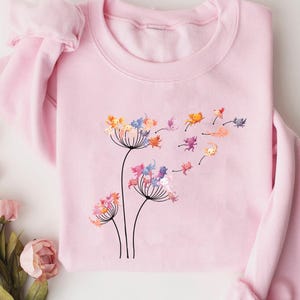 May include: A pale pink sweatshirt with a floral design and small, colourful creatures floating away from the flower heads. The long-sleeved top has a crew neck and a soft appearance.