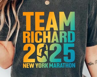 Może przedstawiać: Ciemnoszara koszulka z napisem "TEAM RICHARD 2025 NEW YORK MARATHON" w gradientowym kolorze pomarańczowym, żółtym i niebiesko-zielonym. Numer 2 zawiera sylwetkę biegacza. Styl casual.
