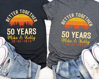 Puede incluir: Dos camisetas, una negra y otra gris, con el texto "BETTER TOGETHER" sobre un gráfico de puesta de sol y "50 YEARS" debajo. Los nombres "Mike & Kelly" y una fecha están impresos debajo. También hay vaqueros y zapatillas blancas en la imagen.