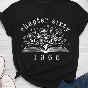 May include: Black t-shirt with a white graphic of an open book with flowers and the text "chapter sixty 1965". The shirt is paired with white sneakers and blue jeans.