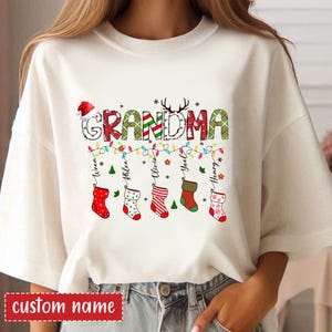 以下が含まれることがあります： クリスマスをテーマにしたデザインのクリーム色のTシャツ。「GRANDMA」の文字が、サンタの帽子とトナカイの角で飾られたお祝いの文字で表示されています。下には、クリスマスの靴下とともに、パーソナライズされた名前がライトの列から吊り下げられています。