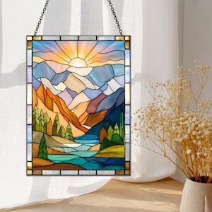 May include: Stained glass panel depicting a mountain landscape with a setting sun. The artwork features a vibrant color palette of blues, oranges, and greens, with a black metal frame and hanging chain. The design includes mountains, trees, and a body of water.
