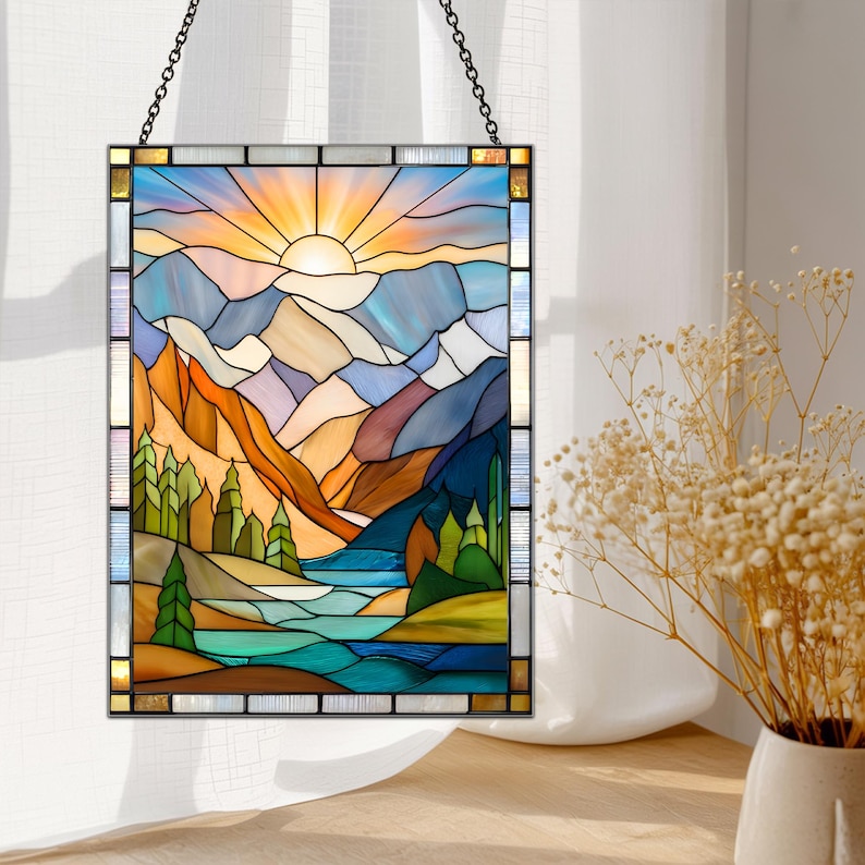 May include: Stained glass panel depicting a mountain landscape with a setting sun. The artwork features a vibrant color palette of blues, oranges, and greens, with a black metal frame and hanging chain. The design includes mountains, trees, and a body of water.