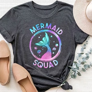 Pu&ograve; includere: Una maglietta grigia scura con una grafica di coda di sirena colorata e il testo "Mermaid Squad".