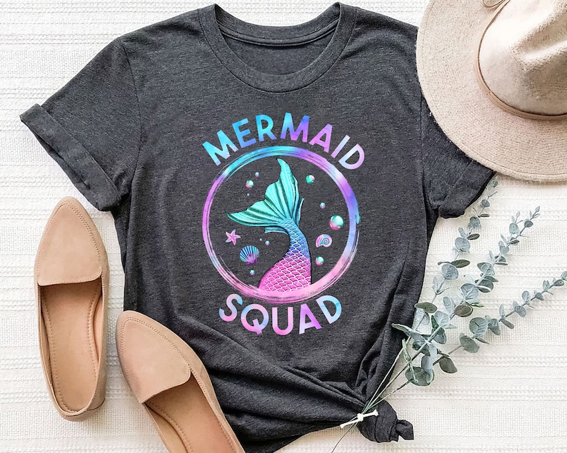 Pu&ograve; includere: Una maglietta grigia scura con una grafica di coda di sirena colorata e il testo "Mermaid Squad".