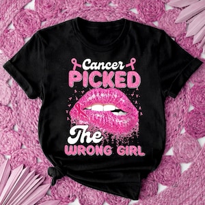 Puede incluir: Camiseta negra con un diseño de labios de purpurina rosa y el texto "Cancer Picked The Wrong Girl" con detalles de cinta rosa.