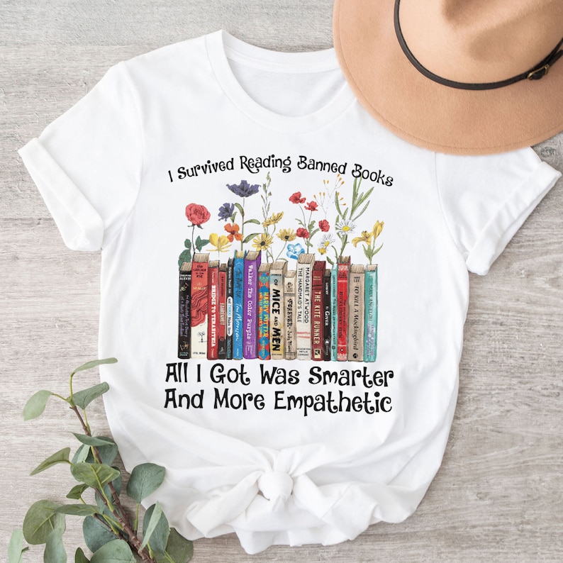 Op de afbeelding: Wit T-shirt met een afbeelding van een stapel boeken met bloemen die eruit groeien. De tekst "I Survived Reading Banned Books" staat boven de boeken en de tekst "All I Got Was Smarter And More Empathetic" staat onder de boeken.