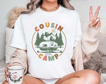 Può includere: Maglietta bianca con una grafica di un camper in una scena di bosco. La grafica è circondata da un cerchio con il testo "COUSIN CAMP" in lettere colorate. L'anno "2024" è stampato sotto il camper.