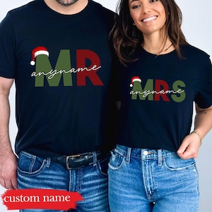 Puede incluir: Dos camisetas negras con temática navideña. Las camisetas dicen "MR" y "MRS" con un gorro de Papá Noel encima de las letras. El texto "anyname" está escrito debajo de las letras en cada camiseta.