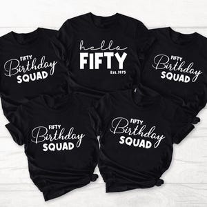 May include: Five black t-shirts with white text. Four shirts read "FIFTY Birthday SQUAD" in a script font. The fifth shirt says "hello FIFTY Est.1975" in a bold font. The shirts are short-sleeved.