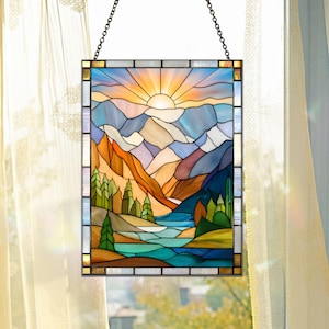 May include: Stained glass panel depicting a mountain landscape with a setting sun. The artwork features a vibrant color palette of blues, oranges, and greens, with a river flowing through the valley. The panel is framed and suspended by a chain.