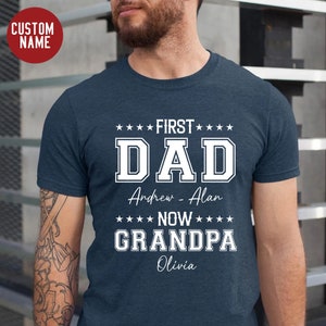 May include: A navy blue t-shirt with white text that reads "First Dad Andrew - Alan Now Grandpa Olivia".