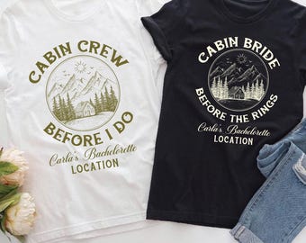 May include: Two white and black t-shirts with a cabin and mountain graphic. The white shirt says "Cabin Crew Before I Do" and the black shirt says "Cabin Bride Before The Rings". Both shirts have the text "Carla's Bachelorette Location".